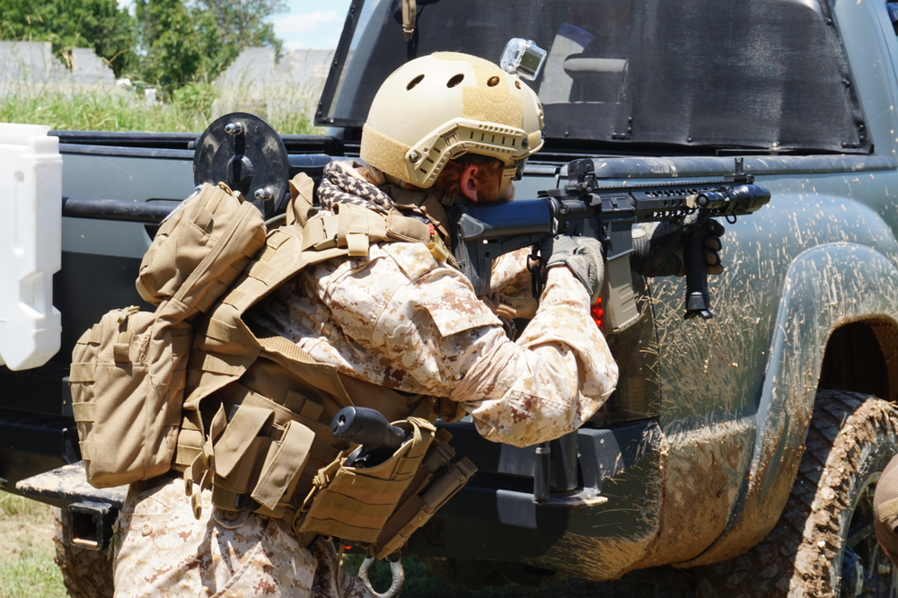 MilSim player taking cover behind truck weilding paintball gun aiming it at somebody