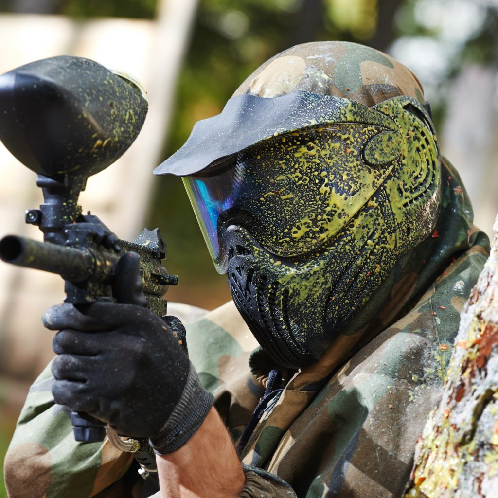 paintball player in protective uniform and mask aiming gun before shooting in summer