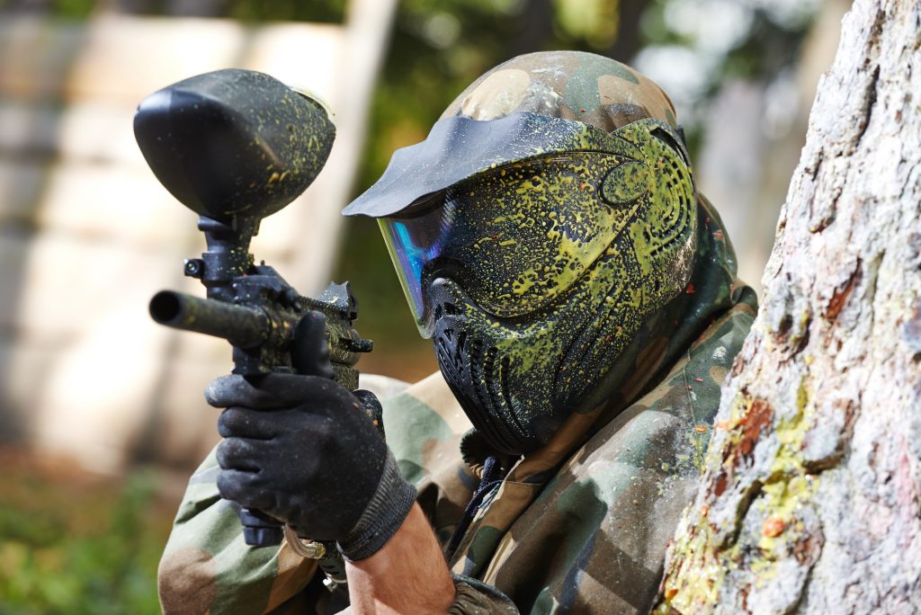 paintball player in protective uniform and mask aiming gun before shooting in summer